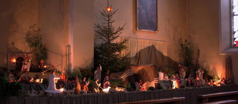 120129krippe_sigdorf002.jpg - Die Weihnachtskrippe orientiert sich an der Bibel. In der Mitte: Die "Krippe" , die Menschwerdung Gottes, in einem Zelt, das über den Resten des Hauses Israel errichtet ist.Vom Kind in der Krippe geht eine Linie über das Kreuz im Zelt hinauf zum Lebensbaum und dem Stern/der Ostersonne: Geburt - Tod - Auferstehung.Links vom Zelt  die Kindheitsgeschichte des Lukas und Motive aus seinem Evangelium, rechts davon die Kindheitsgeschichte das Matthäus mit Motiven aus dem (vorweihnachtlichen) Alten Testament und der (nachösterlichen) Kirche des Neuen Testaments. Alle Darstellungen sind letzlich auf die Mitte, auf Jesus Christus hin ausgerichtet. Der Betrachter ist eingeladen, sich in Situationen und Personen wiederzufinden und sich mit diesen auf den Weg zu machen.